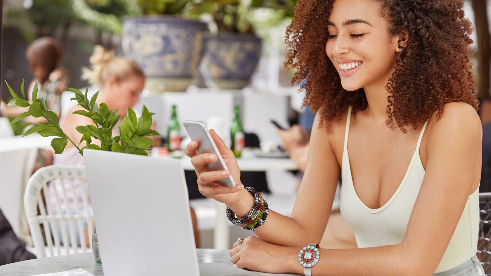 Photo of pleased attractive dark skinned mixed race woman happy to recieve text message from friend, going to make video call on laptop computer, sits against cafe interior. People and lifestyle