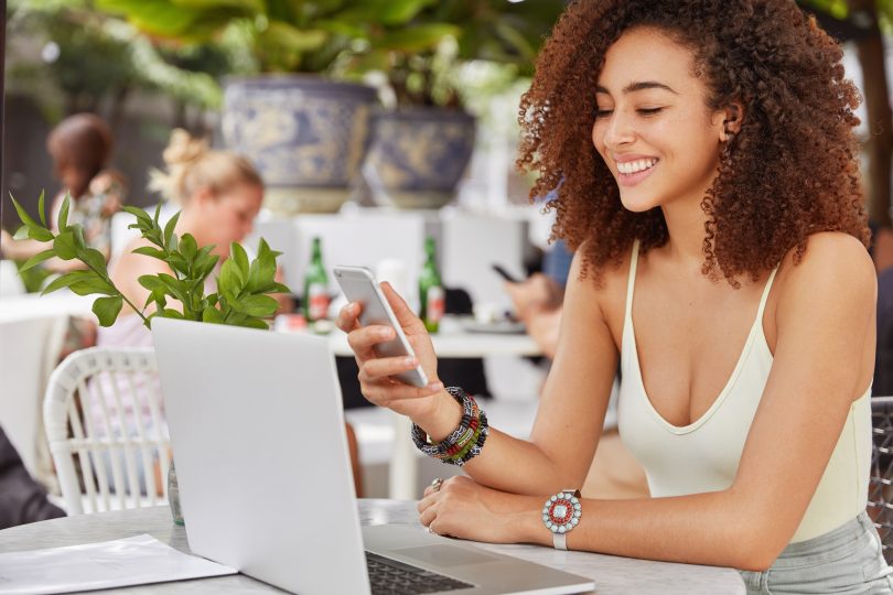 Photo of pleased attractive dark skinned mixed race woman happy to recieve text message from friend, going to make video call on laptop computer, sits against cafe interior. People and lifestyle